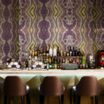 Brown leather bar stools at a marble bar, abstract purple and green wallpaper on wall behind drinks at the bar