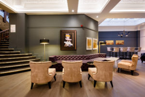 Velvet armchairs and a low leather chesterfield sofa with modern artwork on the wall and sweeping staircase in corner
